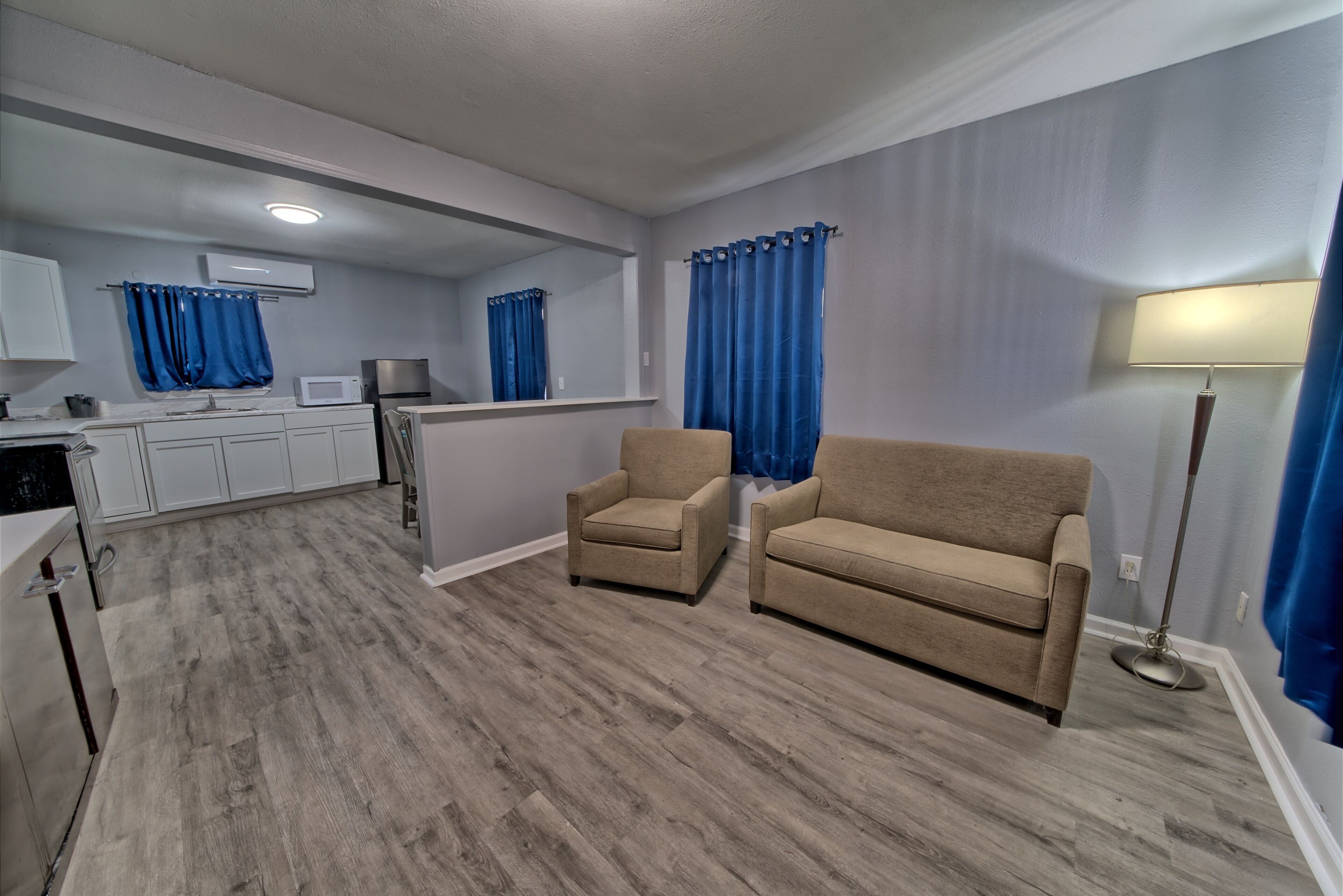 A living room with a beige sofa, a small armchair, a kitchen area with white cabinets, a microwave, and a refrigerator.