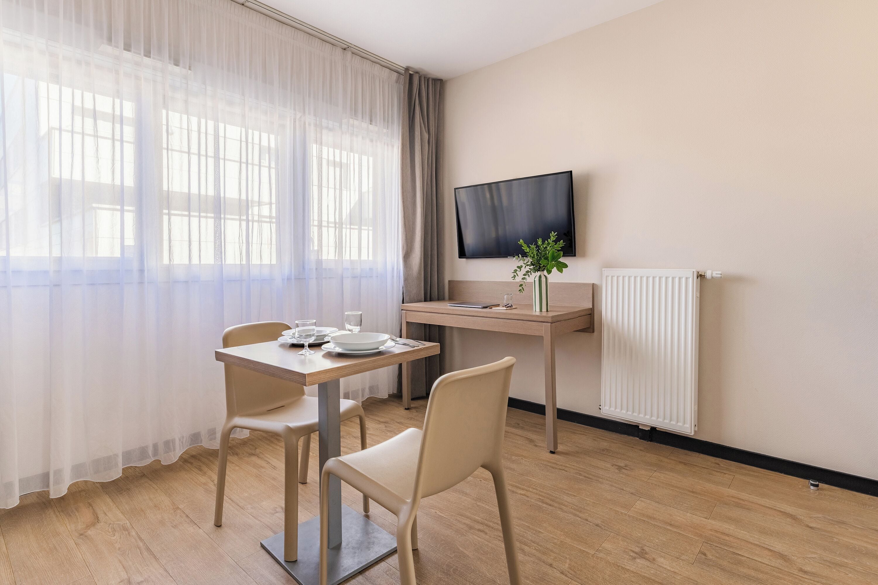 A hotel room with a wooden floor, a dining table with chairs, a TV mounted on the wall, and a radiator.