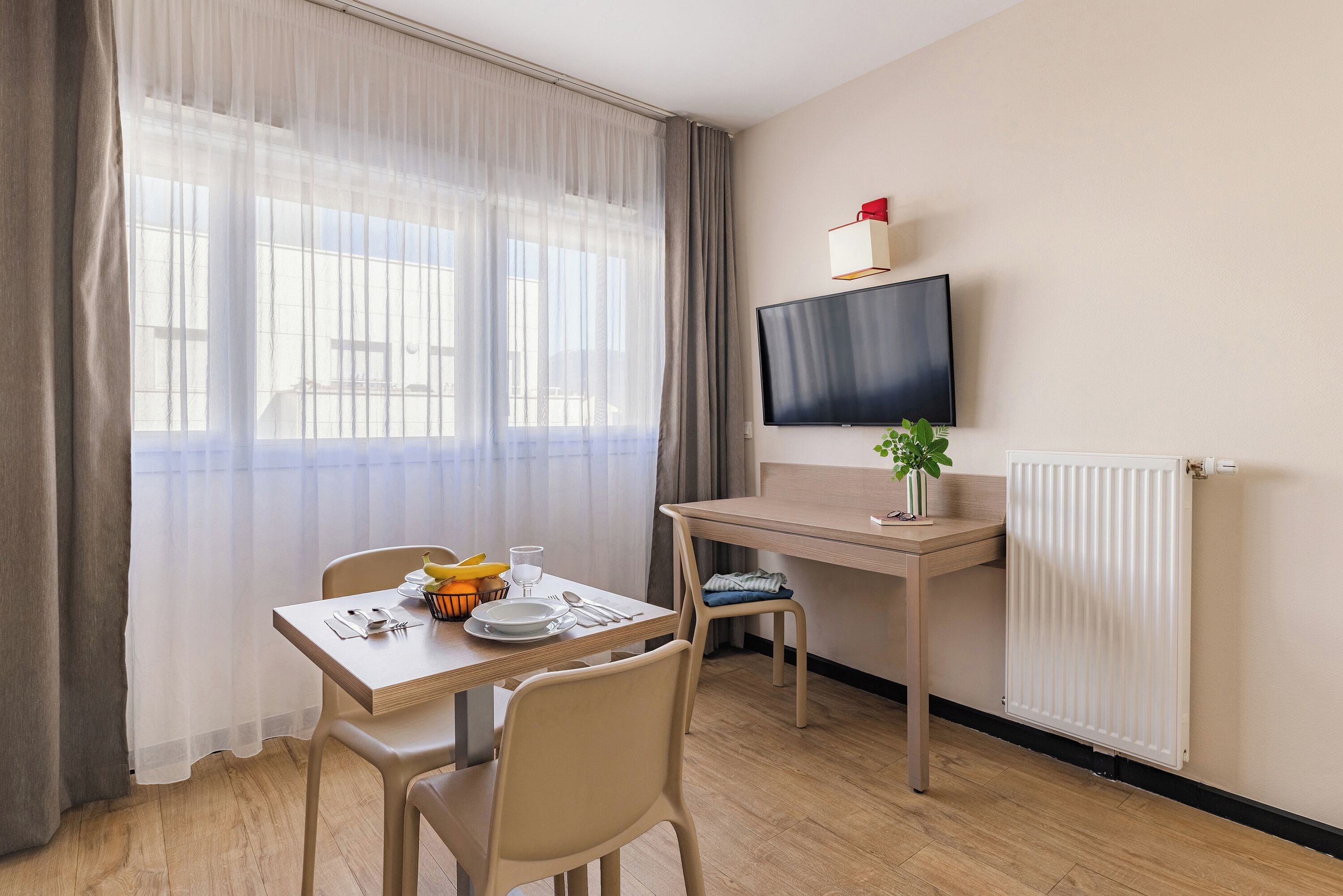 Une chambre d’hôtel équipée d’une table à manger, de chaises, d’un téléviseur fixé au mur, d’un radiateur et d’une fenêtre avec des rideaux légers.
