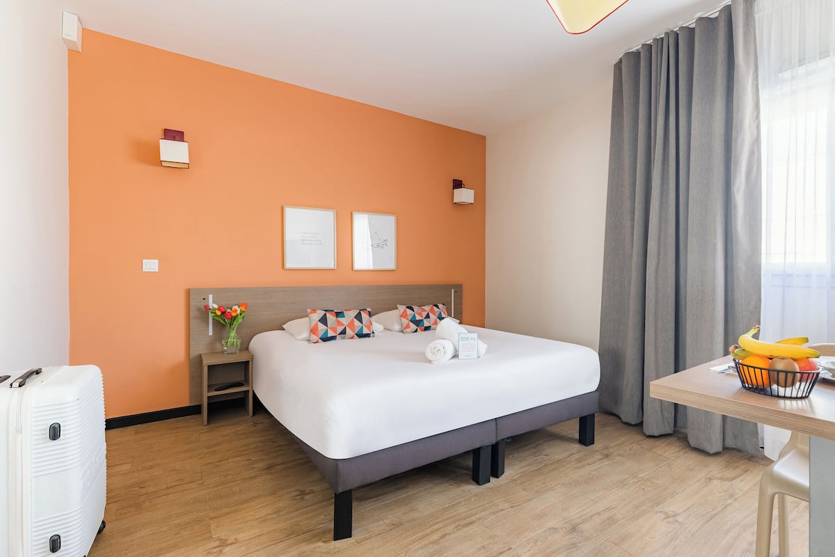 Habitación de hotel con una cama, un escritorio con frutero, una maleta, una mesita de noche con un florero y dos cuadros en la pared.