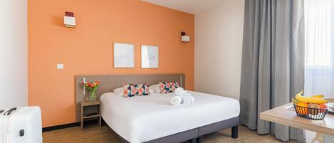 A hotel room with a bed, a desk with a fruit bowl, a suitcase, a nightstand with a vase of flowers, and two framed pictures on the wall.