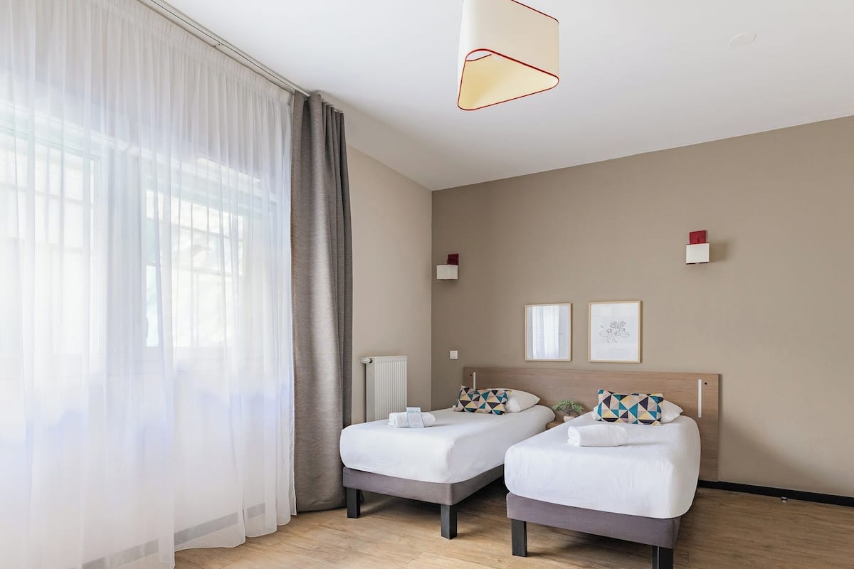 Habitación de hotel con dos camas, un radiador, una ventana con cortinas traslúcidas y una lámpara colgante moderna.