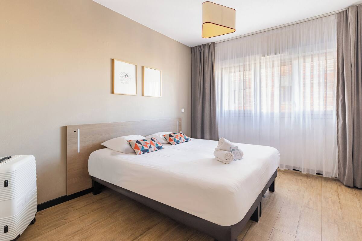 Una cama bien tendida con una colcha blanca y almohadas, una maleta en el suelo y un ventanal con cortinas.