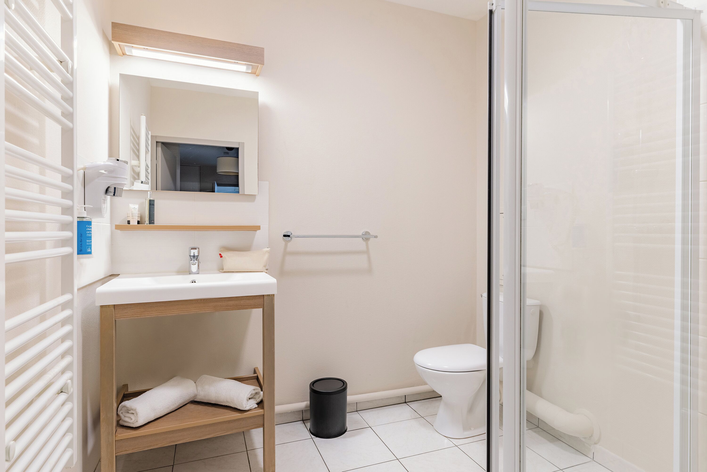 A bathroom with a white sink, mirror, and toilet. There is a towel rack, a trash can, and a shower area.