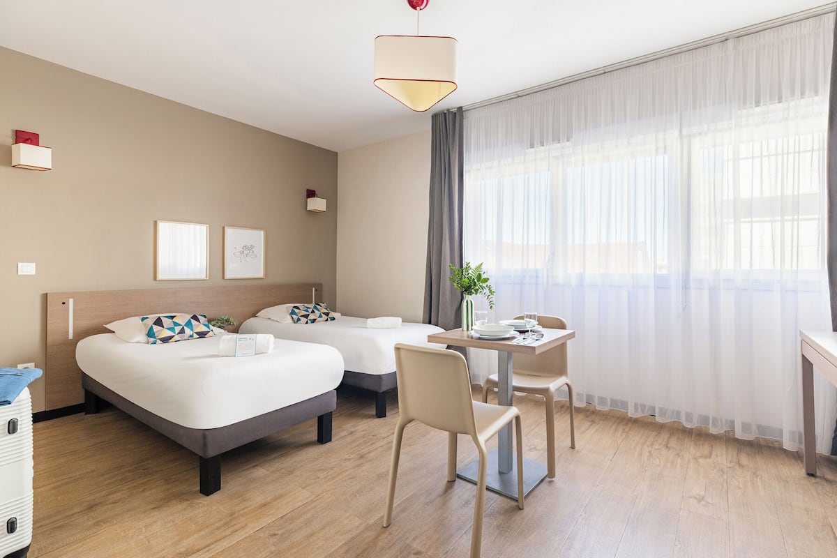 Habitación de hotel con dos camas, una mesa de comedor, sillas, una planta y un gran ventanal con cortinas translúcidas.