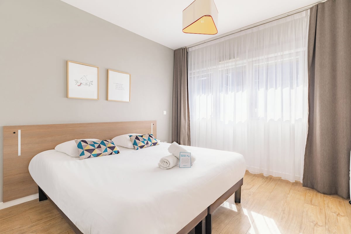Una habitación de hotel con una cama, cabecera, ventana con cortinas y dos cuadros en la pared.