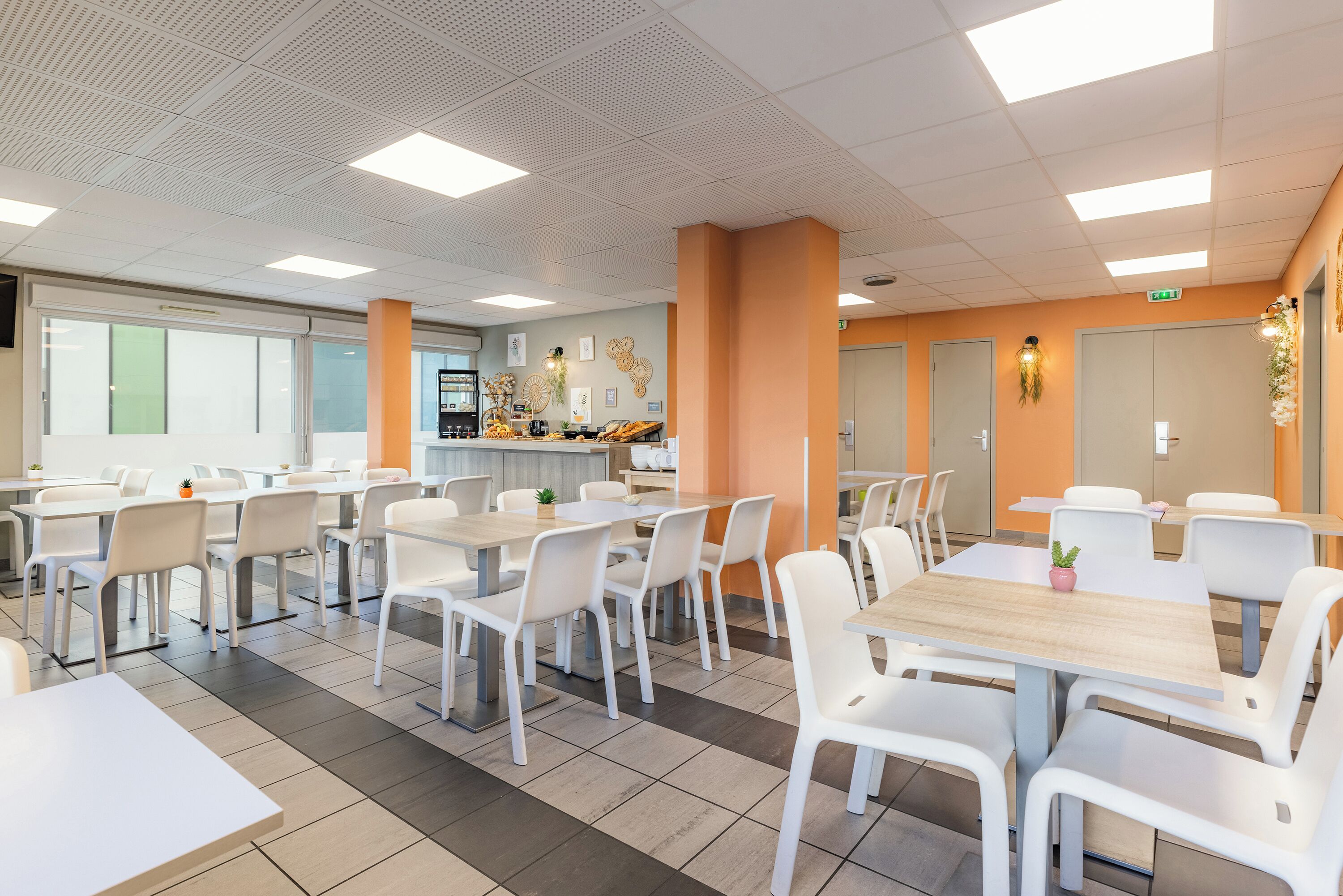 Une cafétéria moderne avec des tables et des chaises blanches, un distributeur de café et un présentoir de fruits.