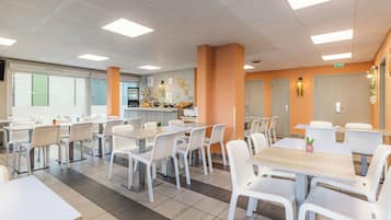 Une cafétéria moderne avec des tables et des chaises blanches, un distributeur de café et un présentoir de fruits.