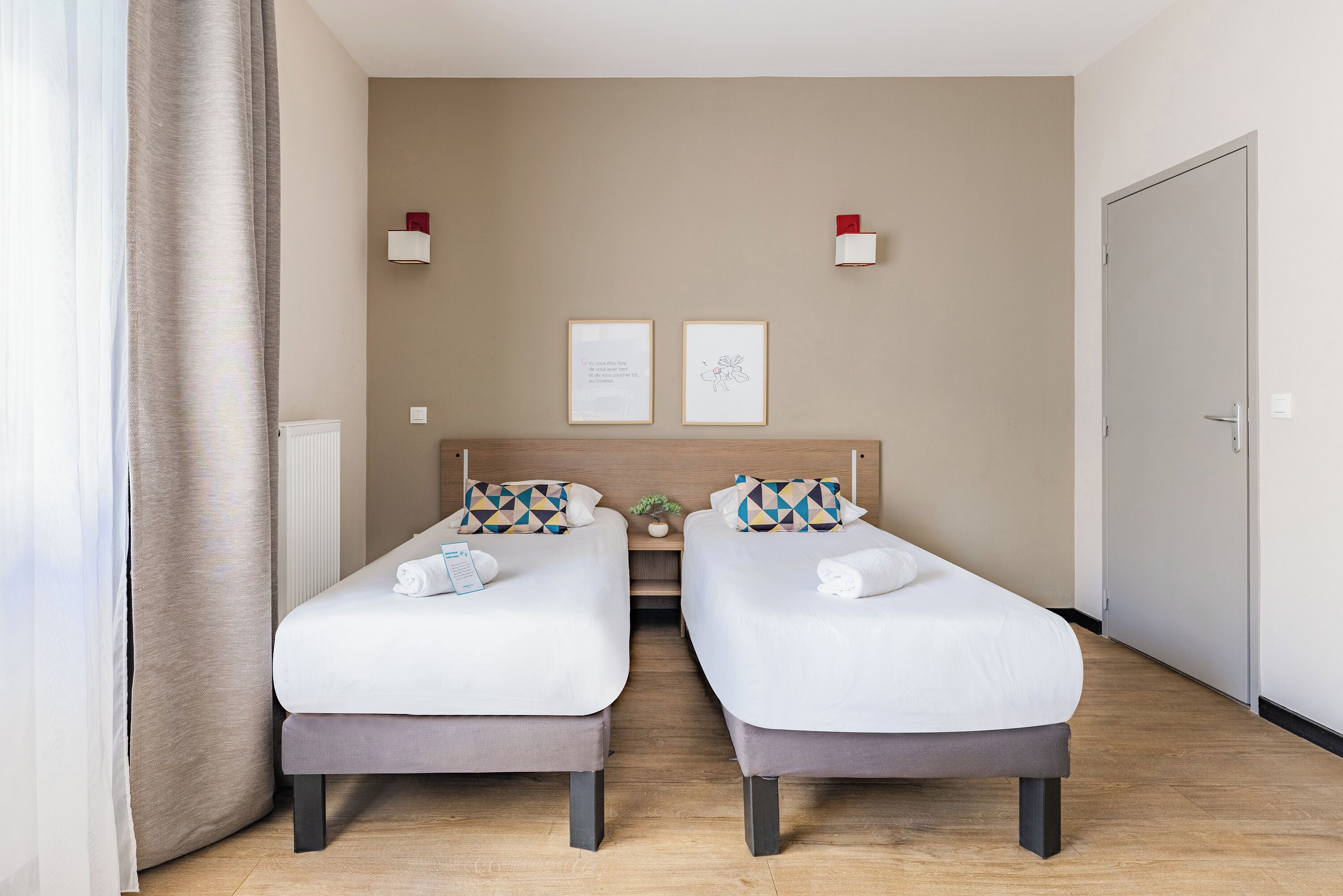 A hotel room with two beds, a small table, a potted plant, and a door.
