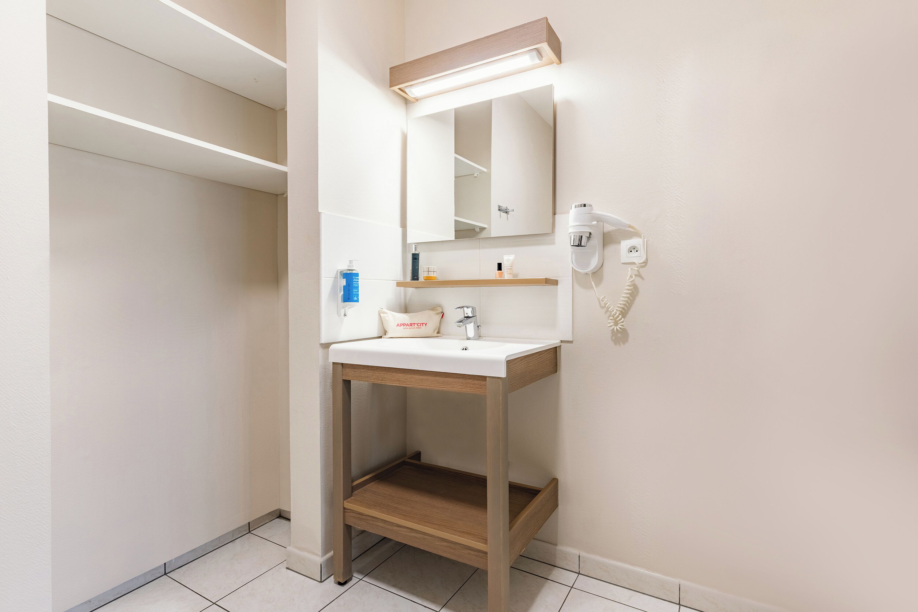 Een badkamer met een wit wastafel, spiegel en haardroger. Boven de wastafel bevindt zich een plank met toiletartikelen.