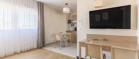 A modern hotel room with a wooden floor, a dining area with a table and chairs, a television mounted on the wall, and a kitchen area in the background.