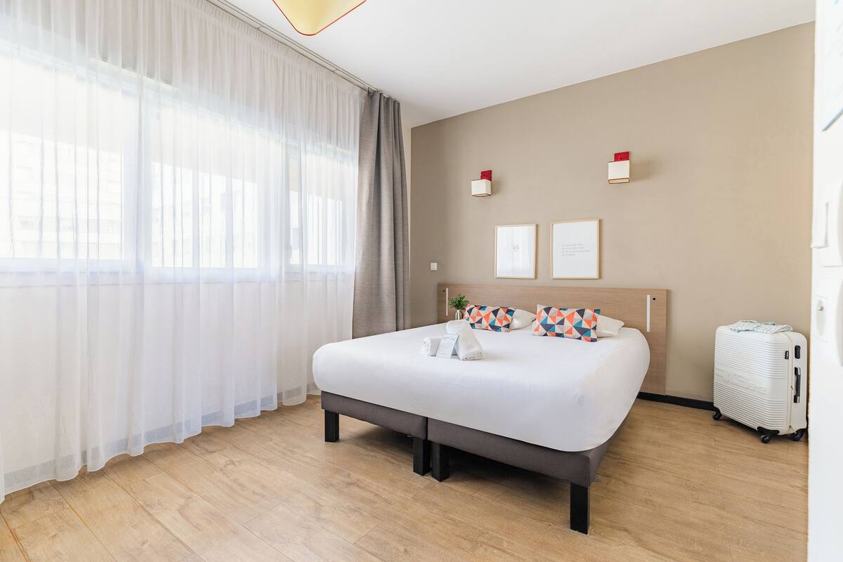Una habitación de hotel con cama, cortinas, cabecera, luces fijadas a la pared y maleta.