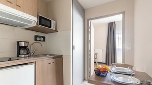 A compact kitchen with a microwave, coffee maker, and a refrigerator. A dining area with a table set for two is visible through an open door. - Appart'City Classic Annemasse Centre-Pays de Genève (Annemasse)