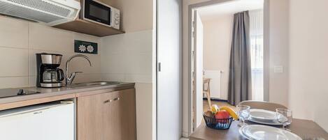 Una cucina compatta con forno a microonde, macchina del caffè e frigorifero. Si intravede una zona pranzo con un tavolo apparecchiato per due persone attraverso una porta aperta.