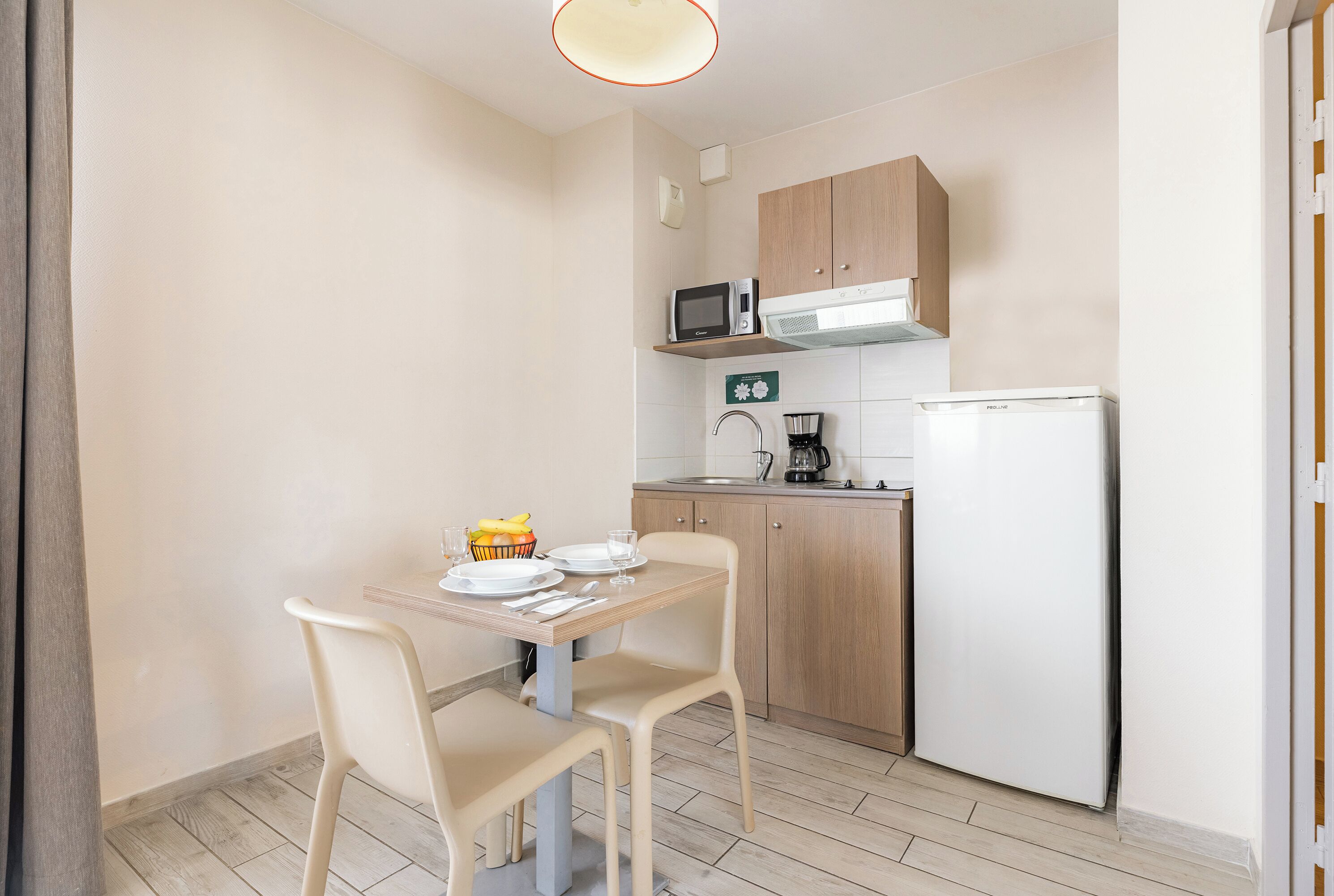 A compact kitchen with a microwave, coffee maker, and refrigerator. There is a dining table with chairs and a small fruit bowl.