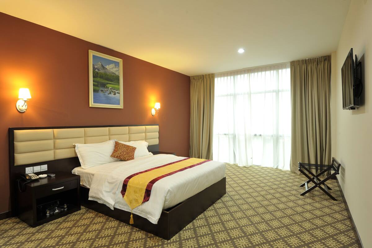 A hotel room with a large bed, a nightstand with a phone, a wall-mounted TV, and a window with curtains.