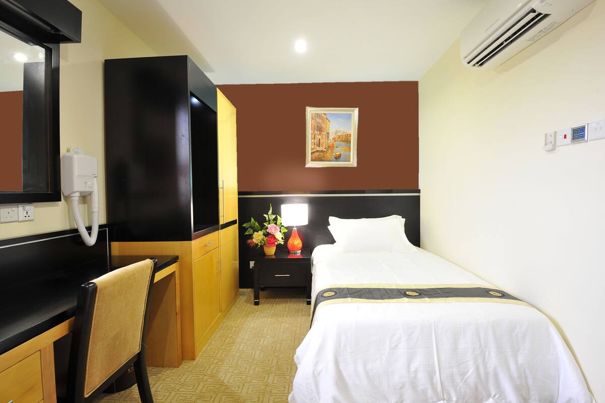 A hotel room with a bed, a desk, a chair, a nightstand with a lamp, and a painting on the wall.