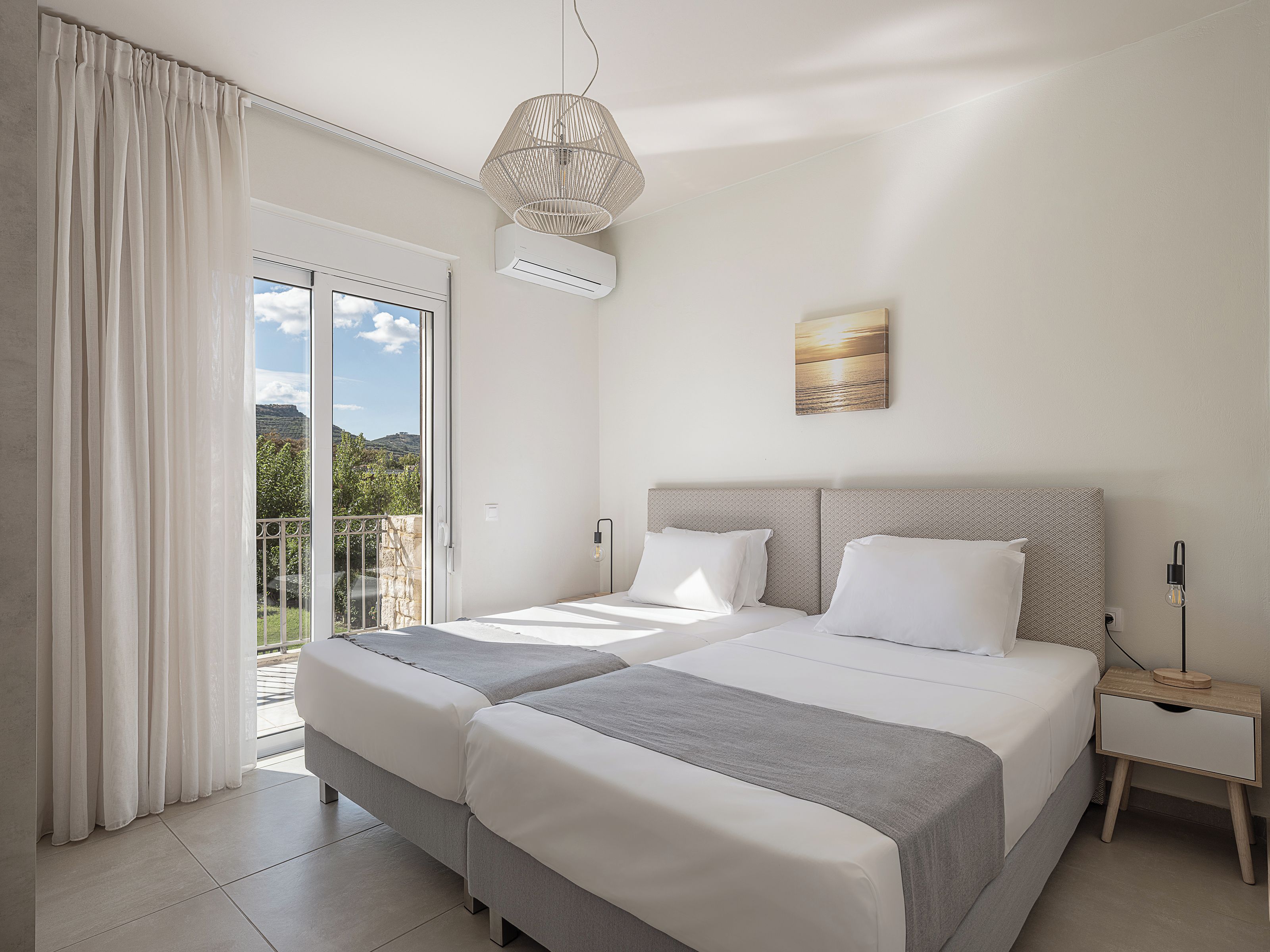 A hotel room with a bed, a nightstand, a wall-mounted air conditioner, a ceiling light fixture, and a view of the outdoors.