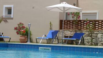 Una piscina amb rajoles blaves, dues gandules blaves, un parasol blanc i una planta en test amb flors roses.