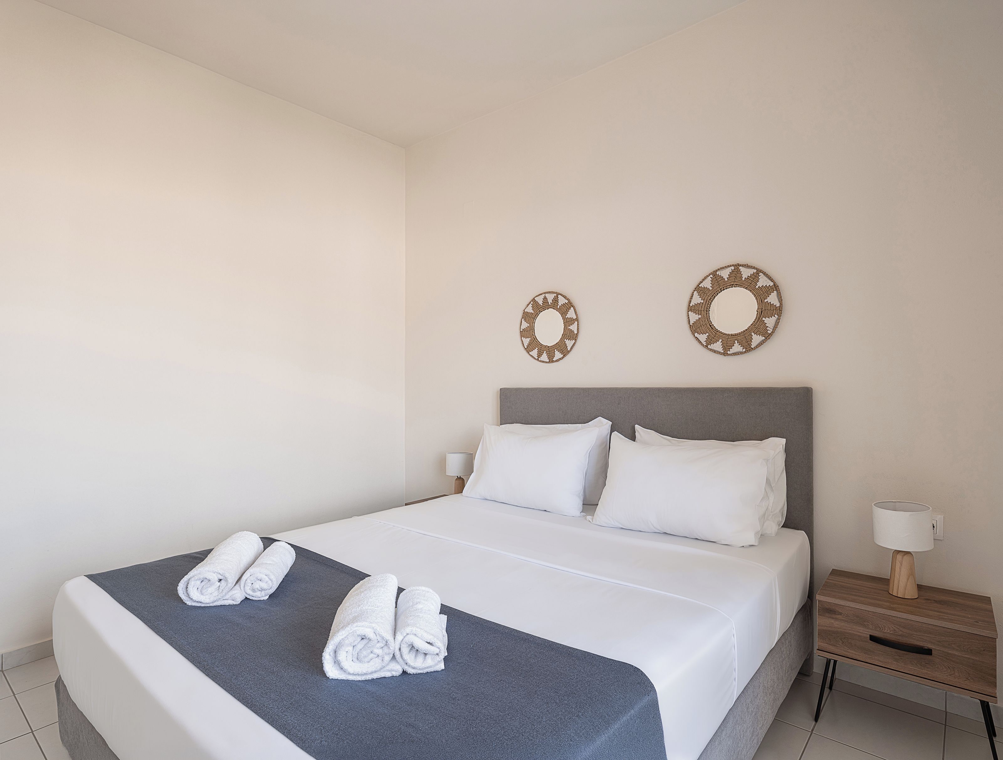 A neatly made bed with white linens and two rolled towels on a grey bedspread.