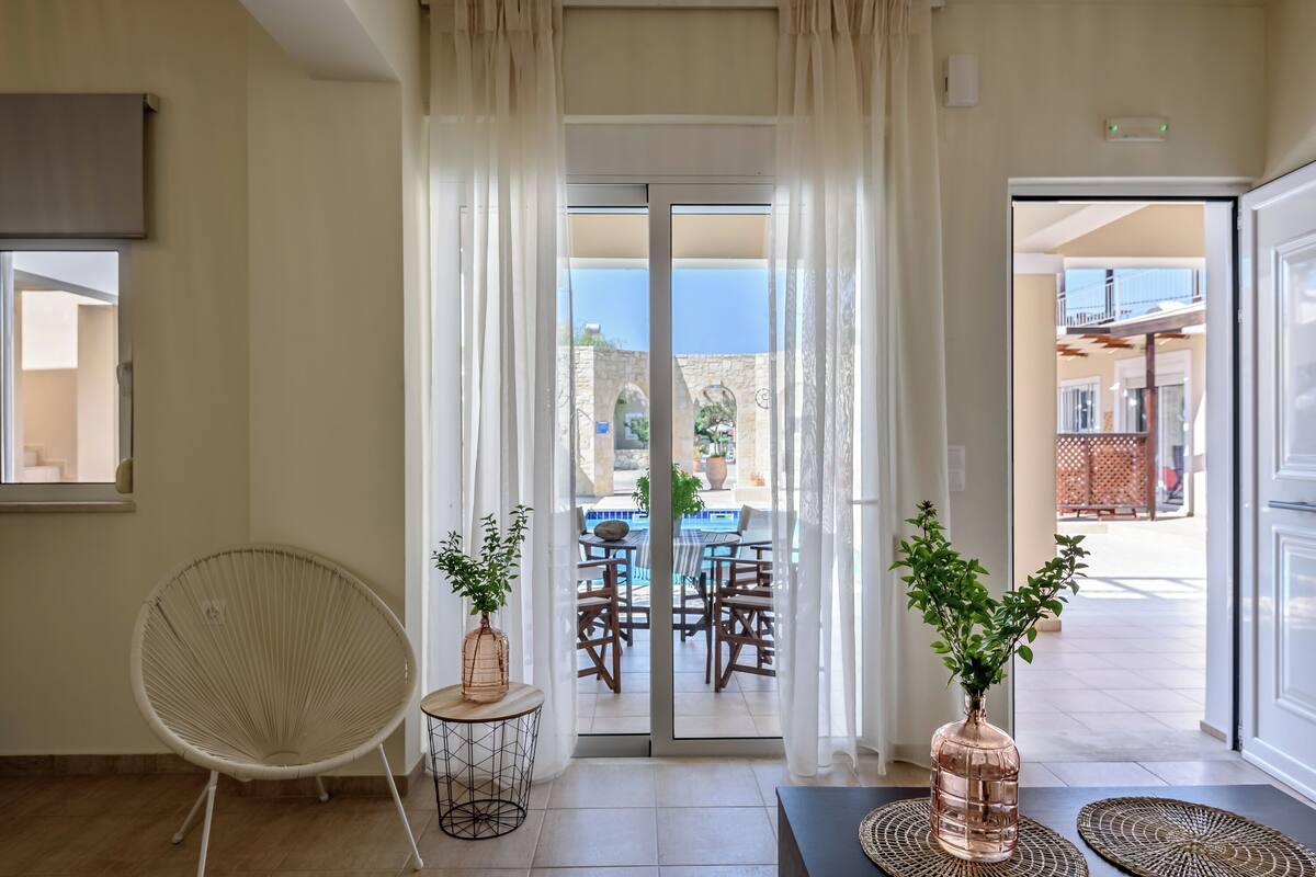 A room with a sliding glass door leading to an outdoor dining area with a pool, a white chair, and two potted plants.