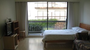 A hotel room with a bed, a TV on a wooden stand, a sofa, and a large window with a view of a building and greenery. - Canada Easy Lodge (Burnaby)