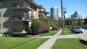 A multi-story residential building with balconies, a sidewalk, parked cars, and modern high-rise buildings in the background. - Canada Easy Lodge (Burnaby)