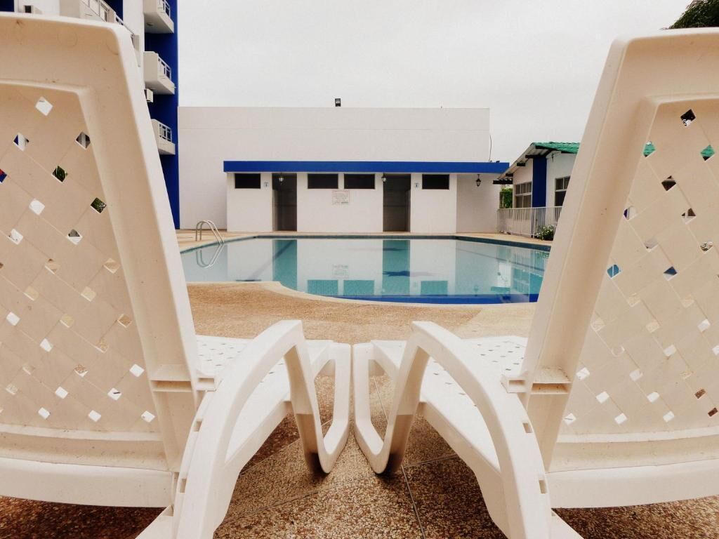 A pool area with white lounge chairs facing a clear pool, a building with blue trim, and a tiled floor.