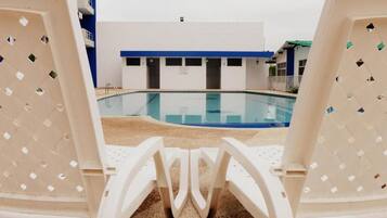 Un área de piscina con sillas de descanso blancas mirando una piscina cristalina, un edificio con detalles azules y un suelo de baldosas.