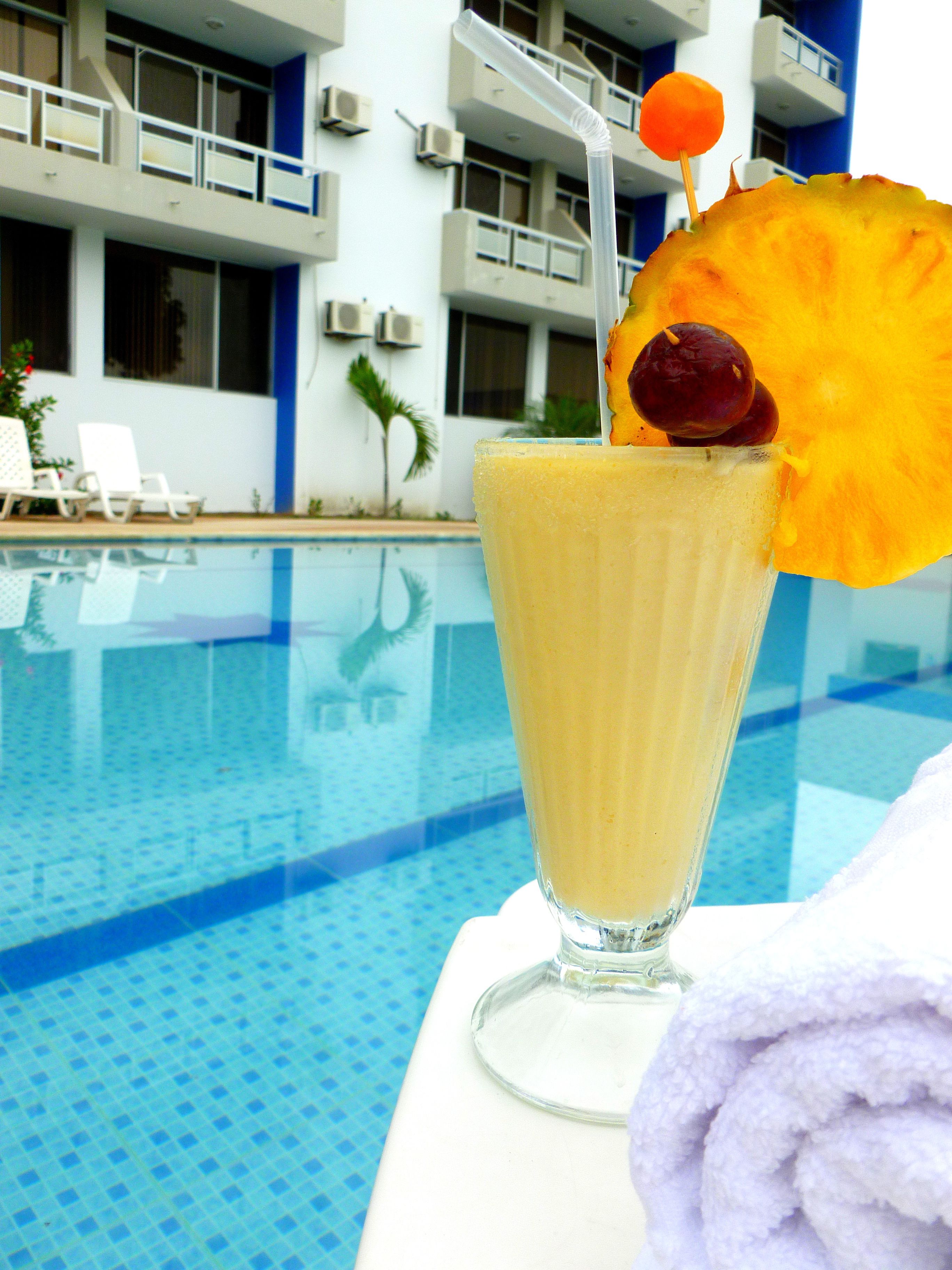 A poolside setting with a tall glass of a yellow beverage, a pineapple slice, and a cherry on a stick.