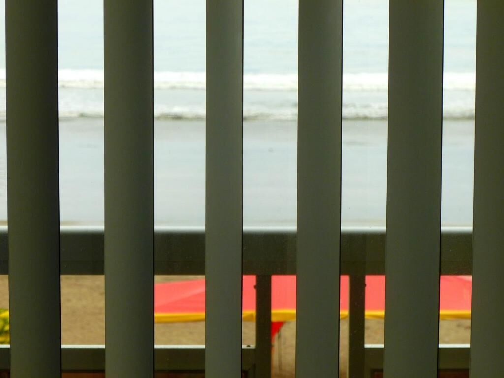 A view through a window with vertical blinds, featuring a beach and a red and yellow object in the foreground.