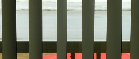 A view through a window with vertical blinds, featuring a beach and a red and yellow object in the foreground.