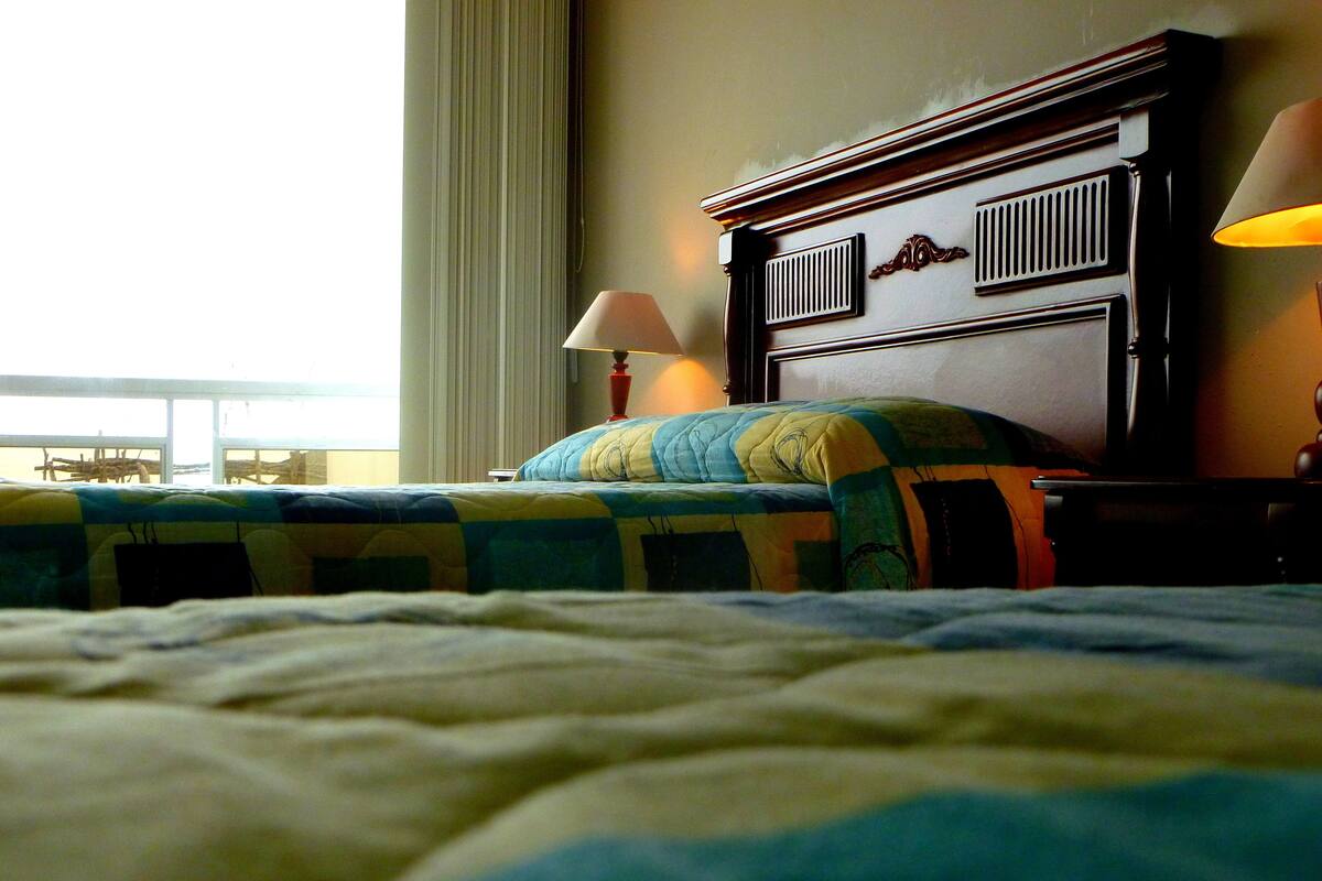 A hotel room with a bed, a nightstand with a lamp, and a view of the beach through the window.
