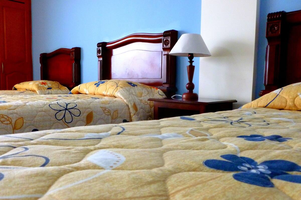 A hotel room with two beds, each with floral-patterned bedding. There is a wooden nightstand with a lamp between the beds.