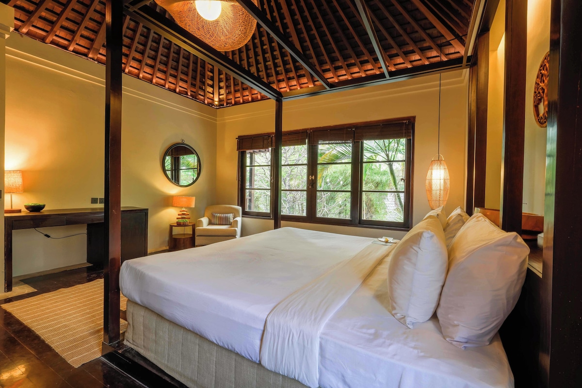A hotel room with a large bed, a desk, a chair, and a window with a view of greenery.
