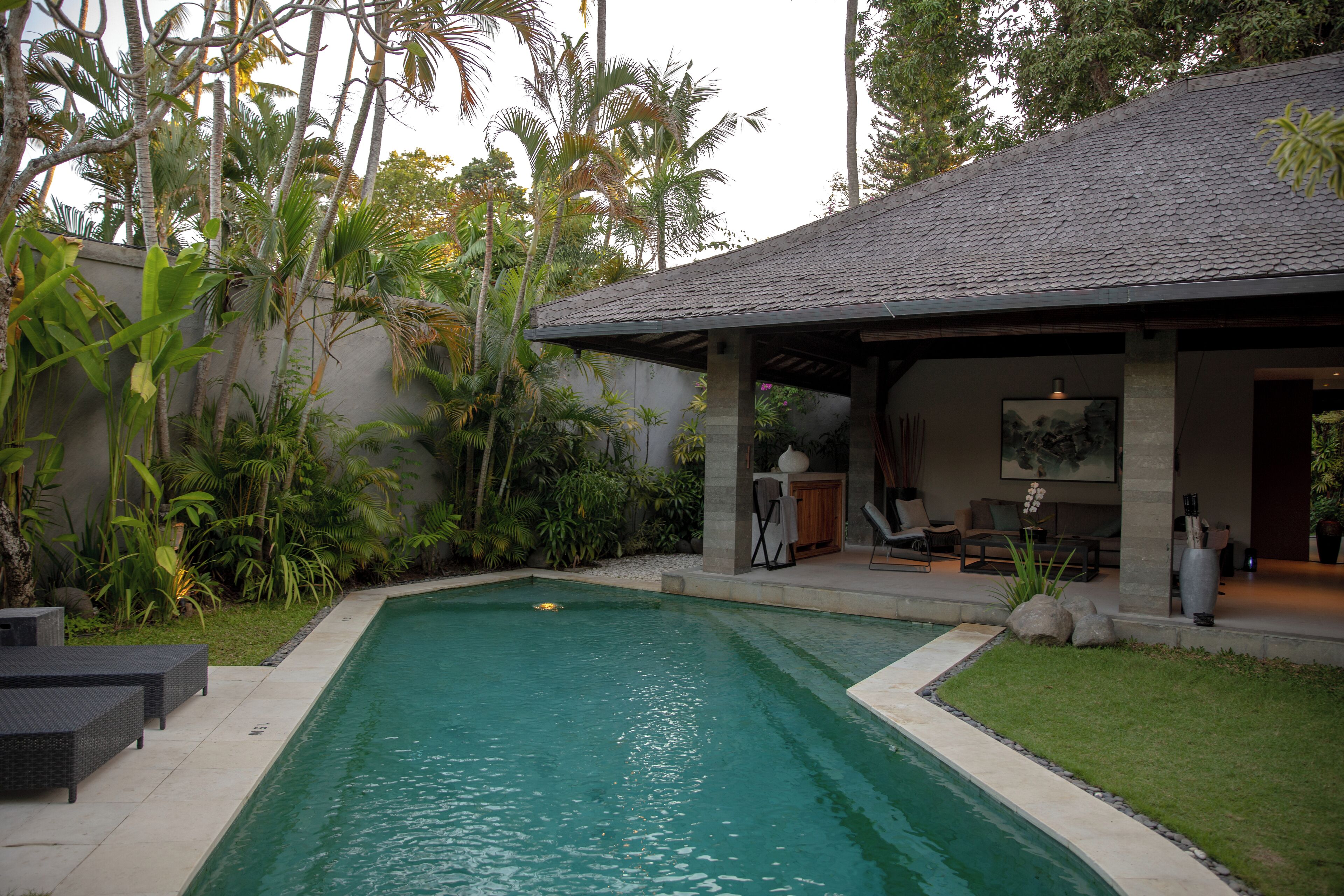 Piscina com área de estar coberta, rodeada por vegetação exuberante e um telhado de pedra.