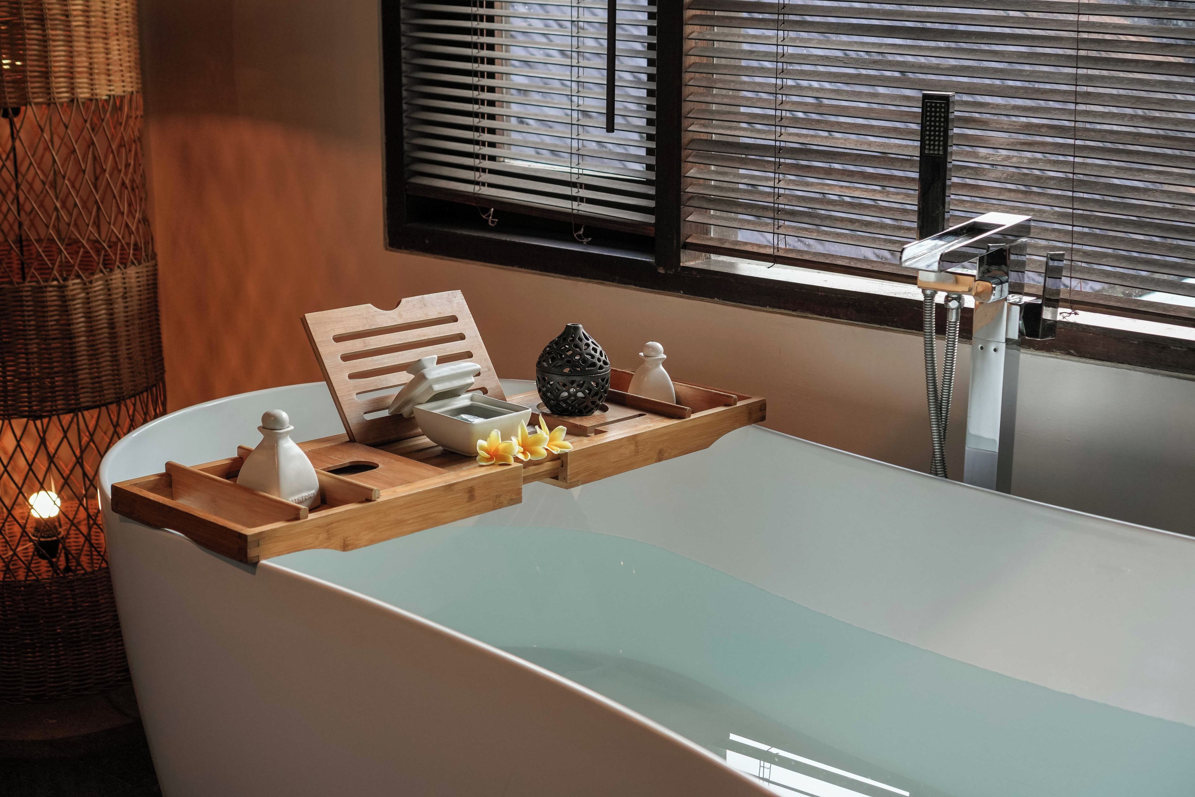 A bathroom with a bathtub, a wooden tray with toiletries, a window with blinds, and a shower area.