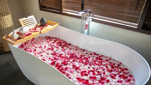 Une baignoire remplie de pétales de rose, un plateau en bois avec une bougie et une fenêtre avec des stores.