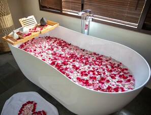 Una bañera llena de pétalos de rosa, una bandeja de madera con una vela y una ventana con persianas.