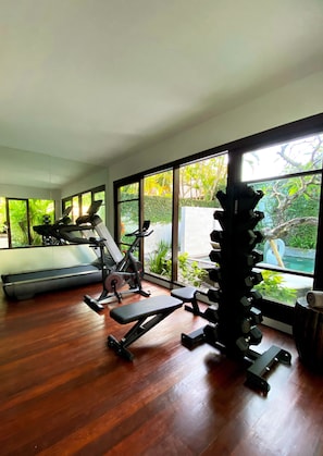 A home gym with exercise equipment, large windows, and a view of greenery. - The Amala Boutique Retreat (Seminyak)