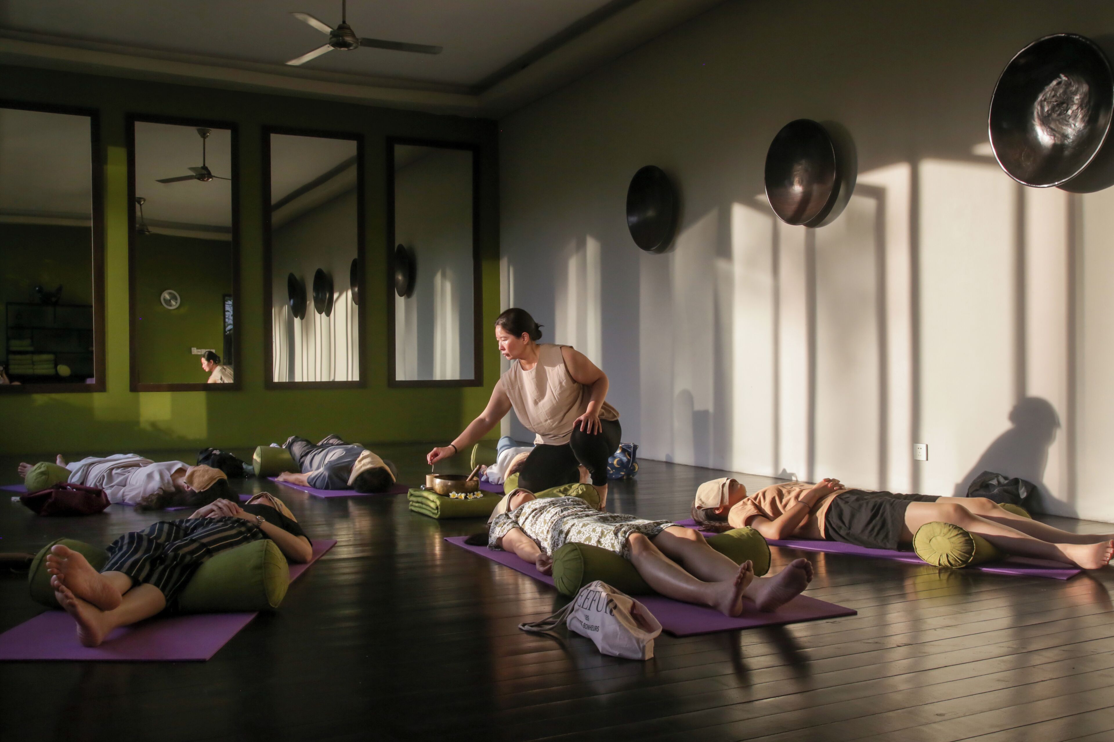 A yoga class in a room with large windows, mirrors, and a ceiling fan.