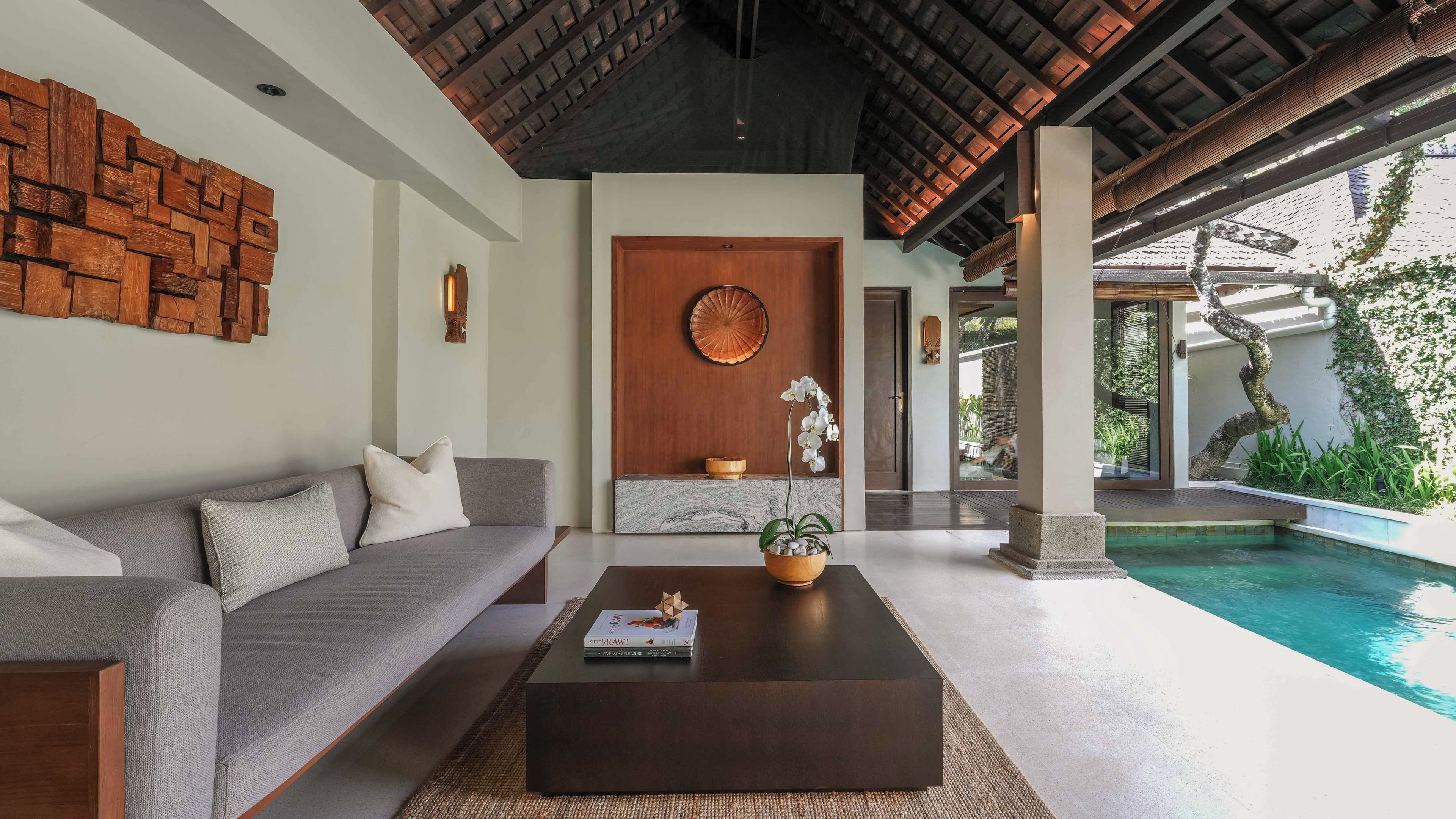Ein modernes Interieur mit grauem Sofa, einem Couchtisch, einer Holzwand und Blick auf einen Swimmingpool.