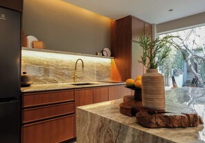 A modern kitchen with a marble countertop, a sink with a gold faucet, wooden cabinets, and a potted plant on the island. - The Amala Boutique Retreat (Seminyak)