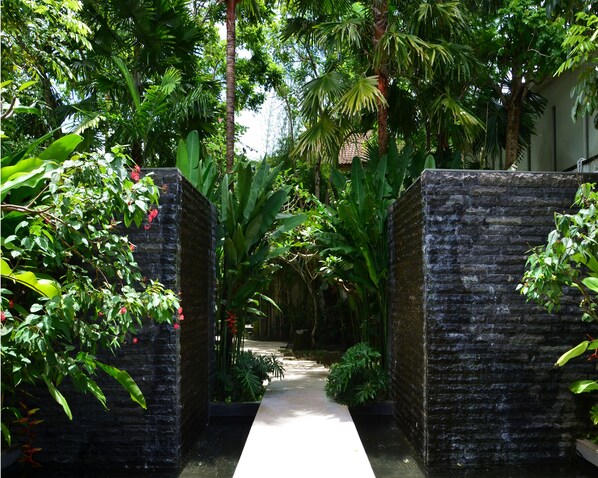 A pathway flanked by stone walls leading through a lush garden with various tropical plants and a clear sky above. - The Amala Boutique Retreat (Seminyak)
