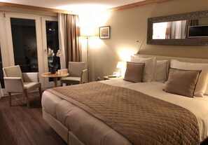 A hotel room with a large bed, a desk with a chair, a small table with a lamp, and a view of the outdoors through a sliding door. - Hotel Albatros Zermatt (Zermatt)