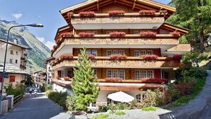 A multi-story wooden building with balconies adorned with red flowers, set against a mountainous backdrop.