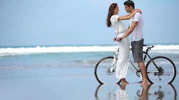 Um casal está em pé na praia com uma bicicleta, ondas do oceano ao fundo.