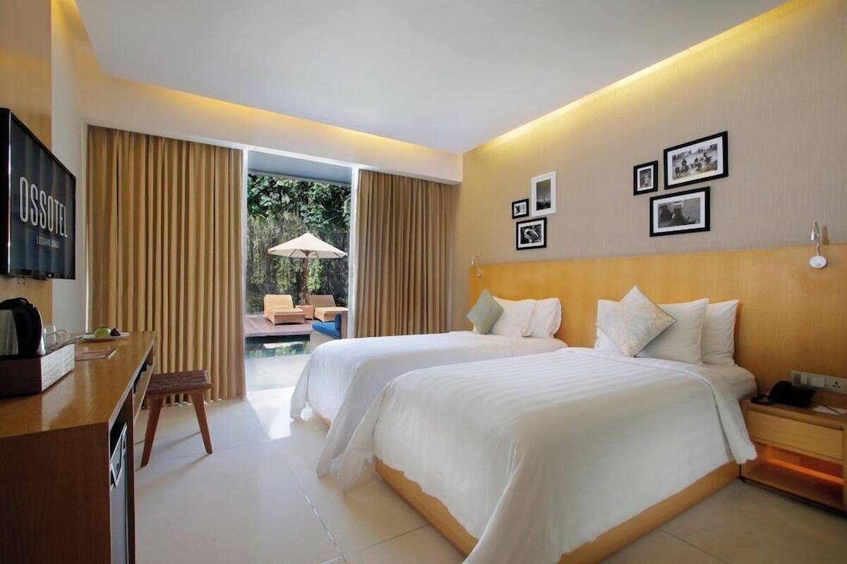 Habitación de hotel con dos camas, un escritorio con televisor, una mesita con sillas y un balcón con cortinas.