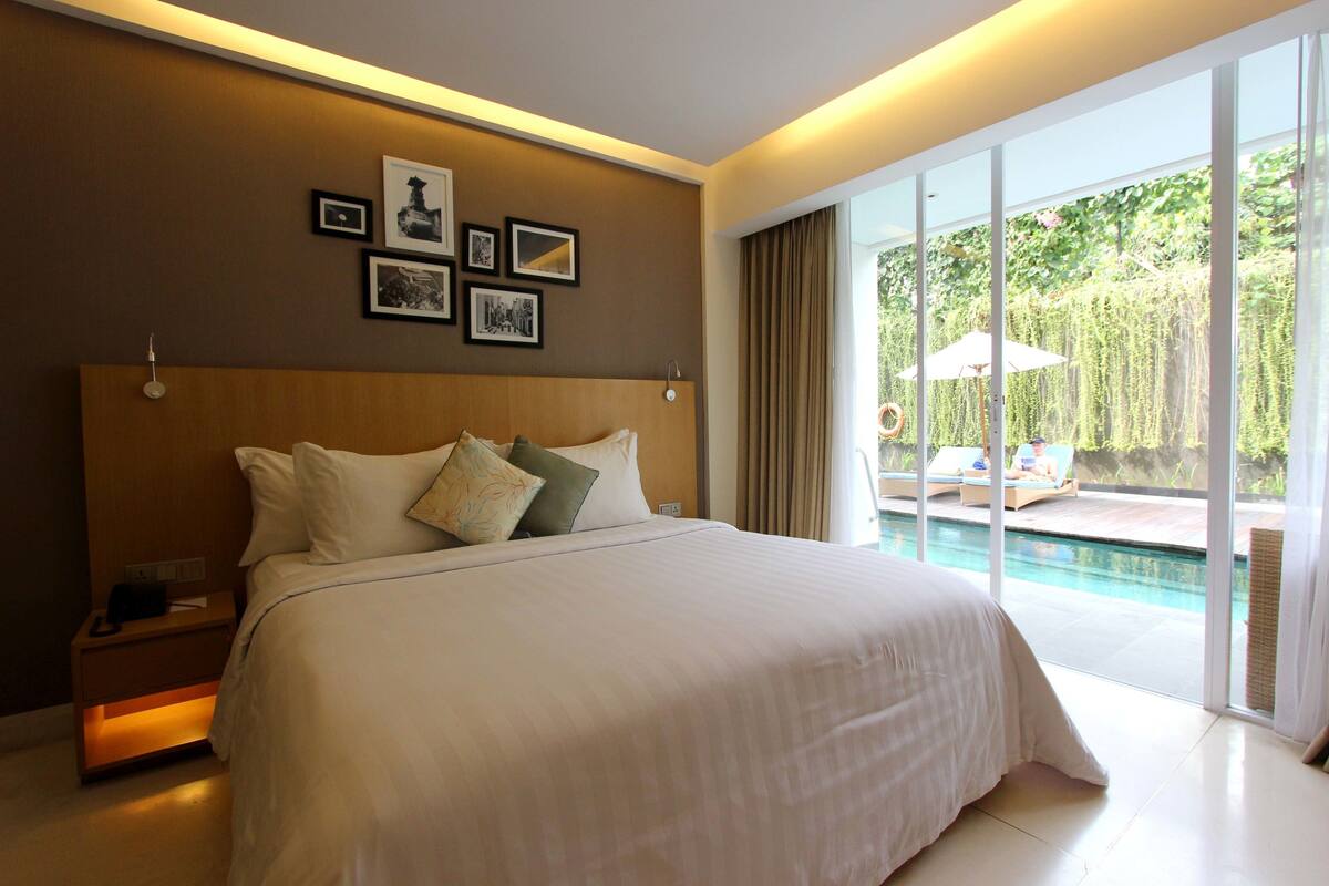 Una cama grande con ropa de cama blanca, cabecera de madera y varios almohadones. En la pared sobre la cama hay fotografías enmarcadas. La habitación tiene un amplio ventanal corredero que da a un espacio exterior con piscina y sillones de descanso.