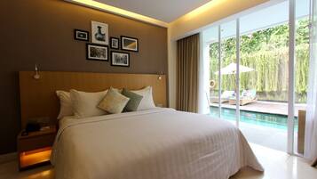 A large bed with white linens, a wooden headboard, and multiple pillows. There are framed pictures on the wall above the bed. The room has a large sliding glass door leading to an outdoor area with a pool and lounge chairs.